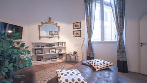 Uma sala de estar com sofá, televisão, estante de livros, espelho e janela com cortinas.