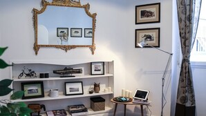 Quarto com parede branca, um espelho grande e ornamentado, quadros, uma estante de livros, uma mesinha com um laptop e um abajur.