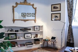 A room with a white wall, a large ornate mirror, framed pictures, a bookshelf, a small table with a laptop, and a floor lamp. - Apartments Florence - Pandolfini Suite (Firenze)