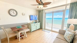 A room with a wooden floor, a dining table with chairs, a television, a ceiling fan, a large window with a view of the sea, and a sofa with a cushion. - Executive Pineapple Escape-Ocean View & Relaxation (Myrtle Beach)