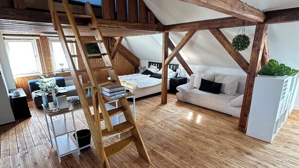 A loft-style room with wooden beams, a wooden ladder, a white sofa, a bed, and a white radiator. - Cozy attic apartment main station/city (Nürnberg)