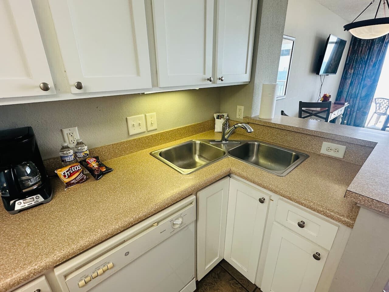 A kitchen with white cabinets, a double sink, a coffee maker, and a TV mounted on the wall.