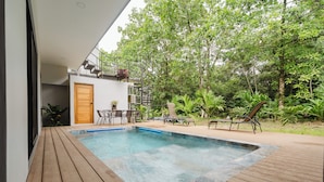 Un área de piscina moderna al aire libre con cubierta de madera, un pequeño espacio de descanso y abundante vegetación.