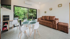 Una sala moderna con un sofá de cuero marrón, una mesa de comedor de cristal con sillas blancas y un ventanal que ofrece vistas a zonas verdes.