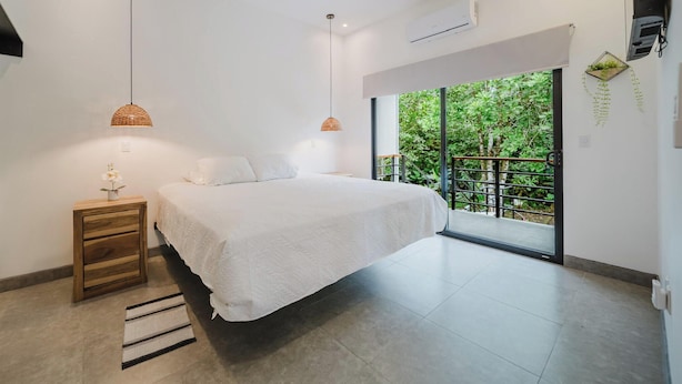 Un dormitorio con una cama, lámparas de noche, una mesita de noche, una ventana con vistas a la vegetación y un aire acondicionado de pared.