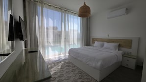 Quarto com cama, mesa de cabeceira, televisão na parede e janela com cortinas leves.