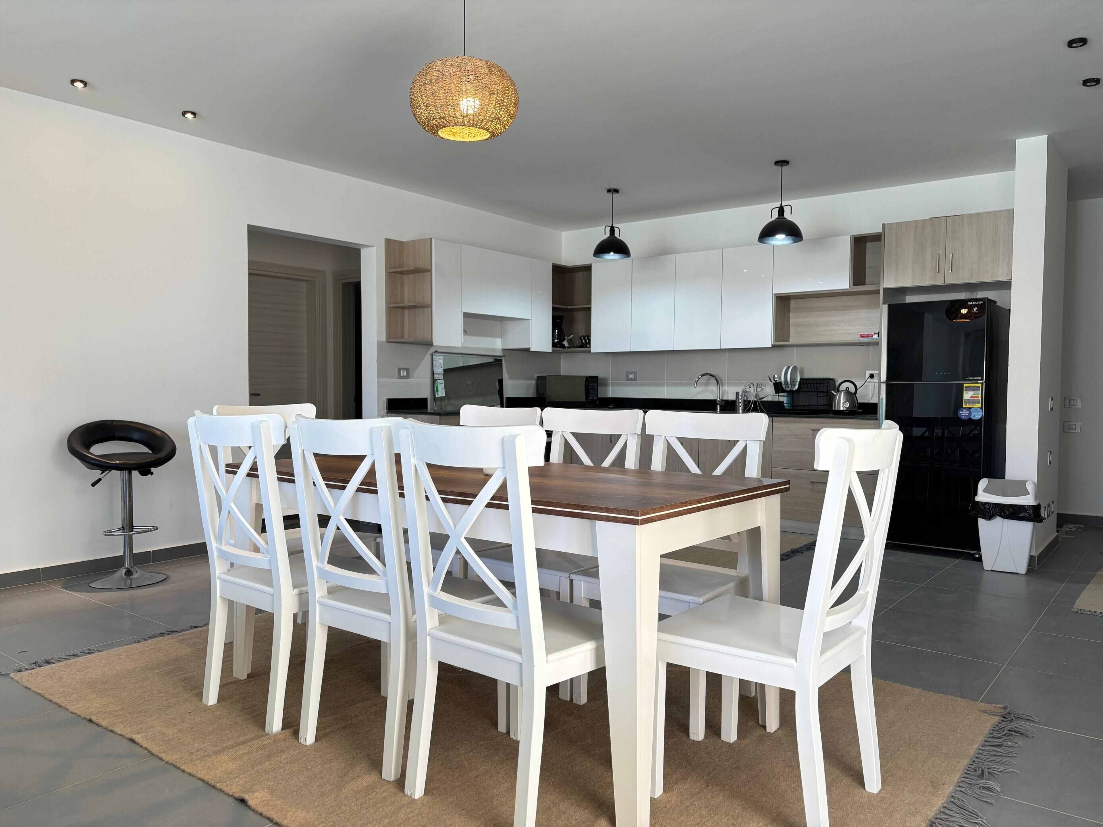 A modern kitchen with white cabinets, a black refrigerator, and a dining area with white chairs and a wooden table.