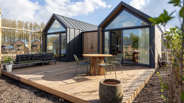 Ein modernes Haus mit großen Glasfenstern, einer Holzterrasse und Gartenmöbeln.