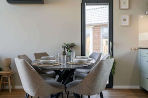 A dining area with a round table, grey upholstered chairs, and a view through a glass door. - Tiny house in Oud Ade | Tuin van Keet - 15 (Oud Ade)