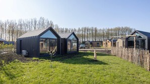A modern, open-plan house with large glass windows, a wooden deck, and a fenced yard. - Tiny house in Oud Ade | Tuin van Keet - 15 (Oud Ade)