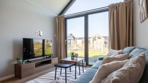 A modern living room with a flat-screen TV, a sofa, a coffee table, and a large window with curtains. - Tiny house in Oud Ade | Tuin van Keet - 15 (Oud Ade)