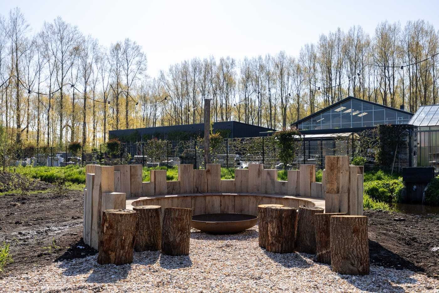 Een rond zitgedeelte van hout met een vuurkorf, omgeven door een grindpad, met een gebouw en bomen op de achtergrond.