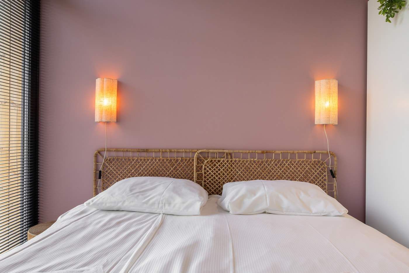 Una cama con ropa blanca, un cabecero tejido y dos lámparas fijadas a la pared.