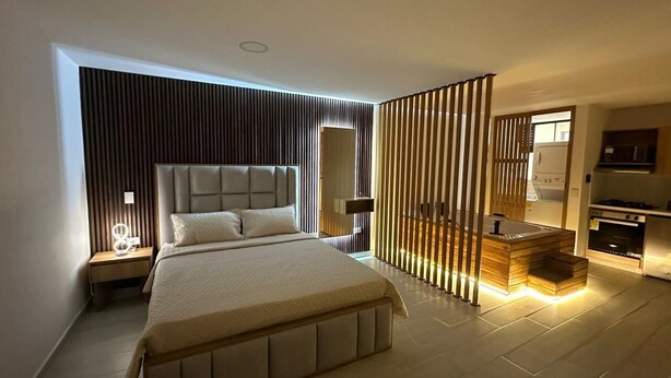 A modern bedroom with a large bed, bedside tables, a built-in bench, and a kitchen area with a stove. - Apartamento con Jacuzzi Parque Guatape Antioquia (Guatape)