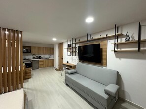 Una sala de estar moderna con un sofá gris, armarios de madera, un televisor y una cocina al fondo.