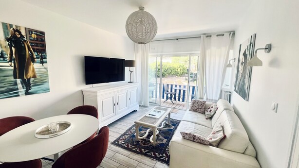 Ein Wohnzimmer mit Fernseher, einer Couch, einem Esstisch, Stühlen und einem Balkon mit Gartenmöbeln.
