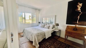 Ein Schlafzimmer mit Bett, Nachttischen, einem Schminktisch und einem Bild, mit Blick durch das Fenster.