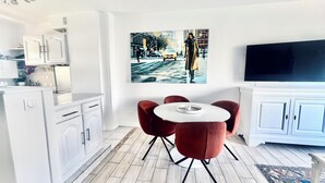 Eine moderne Küche mit weißen Schränken, ein Essbereich mit rundem Tisch und roten Stühlen sowie ein großer Flachbildfernseher an der Wand.