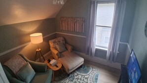 A cozy room with a green armchair, a patterned armchair, a small table with a lamp, a patterned rug, and a window with curtains.