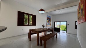 A spacious room with a wooden dining table, benches, a hanging pendant light, a window with shutters, and a colorful painting on the wall. - Vacation home in Porto das Dunas with pool. (Aquiraz)