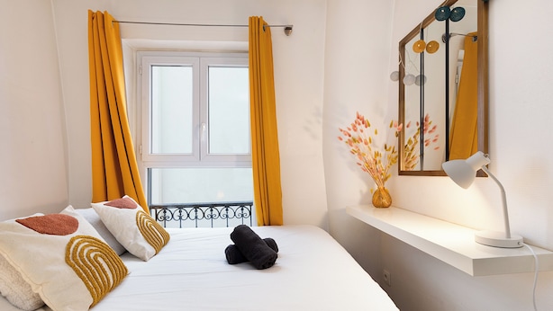 Ein Hotelzimmer mit Bett, einem Fenster mit gelben Vorhängen, einem Nachttisch mit Lampe und einem Spiegel mit Dekorationen.