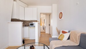 Eine moderne Küche mit weißen Schränken, einer Mikrowelle und einer Kaffeemaschine. Es gibt einen Glastisch mit einer Fernbedienung und einer Obstschale. Ein graues Sofa mit Decke und dekorativen Kissen befindet sich im Vordergrund.