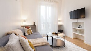 Ein modernes Wohnzimmer mit einer Couch, einem Couchtisch, einem flachbildfernseher an der Wand und einem Fenster mit durchsichtigen Vorhängen.