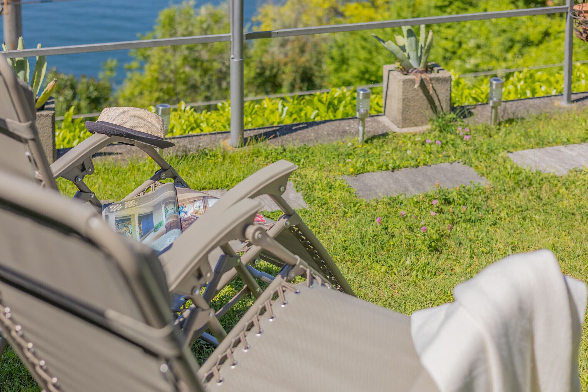 Una tumbona con sombrero y una revista, una barandilla metálica, y vistas a vegetación y una masa de agua.