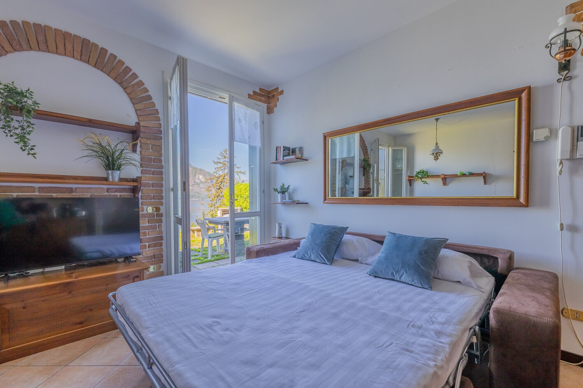 A bedroom with a large bed, a TV, a brick fireplace, and a view of the outdoors.