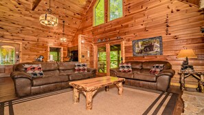 A log cabin interior with two leather sofas, a wooden coffee table, a bear-themed painting, and a chandelier. - Cozy Cabin in Pigeon Forge, Hot tub & Scenic Views (Pigeon Forge)