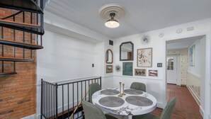 Un coin repas avec une table ronde, quatre chaises, un mur en briques, un escalier et un couloir menant à la cuisine.
