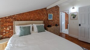 Une chambre avec un mur en briques, un lit avec une literie blanche, une table de chevet, une commode et une porte donnant sur une autre pièce.