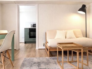 Ein modernes Wohnzimmer mit einer Couch, einem kleinen Holztisch, einer Lampe und einer Küche im Hintergrund.