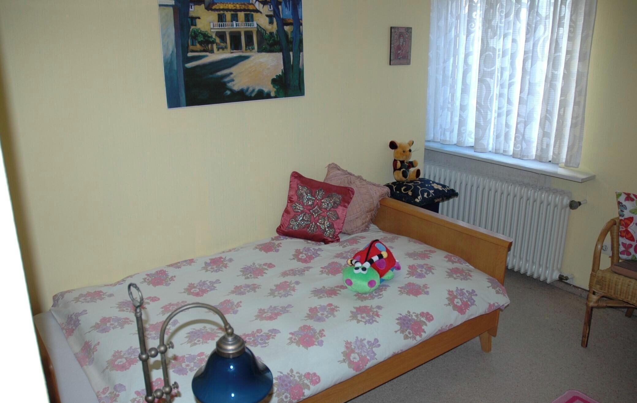Ein Schlafzimmer mit Bett, Nachttisch, Lampe, einem Teddybären und einem Fenster mit Vorhängen.