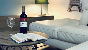 Una habitación moderna con sofá, una mesa con botella y copa de vino, lámpara, estantería y un reloj de pared con diseño de la Torre Eiffel.