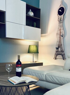 Ein modernes Zimmer mit einer Couch, einem Tisch mit Weinflasche und Glas, einer Lampe, einem Bücherregal und einer Wanduhr mit Eiffelturm-Motiv.