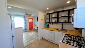 Una cocina compacta con estufa a gas, un lavamanos y una encimera. Se ve una puerta roja que da a otra habitación, y también un ventilador de techo.