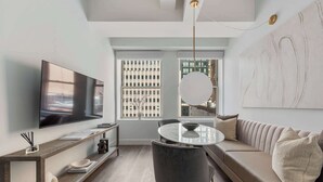 Ein modernes Wohnzimmer mit Flachbildfernseher, einem runden Esstisch, einer Couch und einem großen Fenster mit Stadtblick.