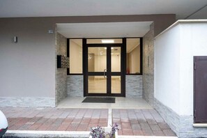 Ein Gebäudeeingang mit Glastür, einer kleinen Außenbestuhlung und einem an der Wand befestigten Schild.