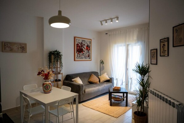 A living room with a sofa, a dining table with chairs, a hanging light fixture, and a window with curtains.