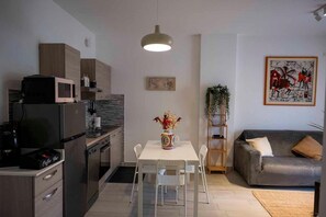 A compact kitchen with modern appliances, a dining table with chairs, a grey sofa, and a painting on the wall.