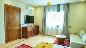 Una habitación con armario de madera, televisión de pantalla plana, una mesita redonda con un jarrón, sofá y un amplio ventanal con cortinas.
