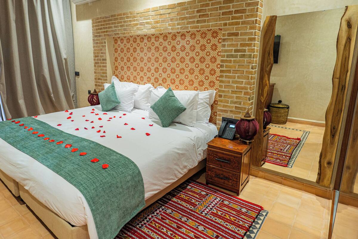 Ein Hotelzimmer mit einem großen Bett, einem Holfnachtkästchen, einem gemusterten Teppich und einer Ziegelwand.