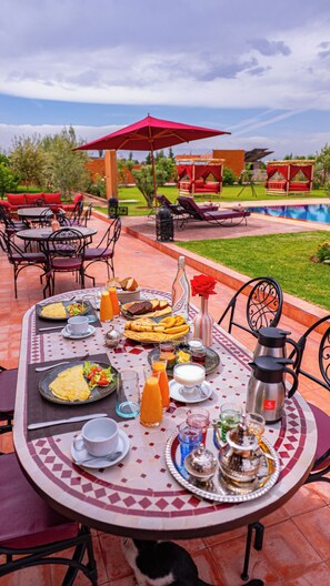 A table set outdoors with a variety of food and drinks, a red umbrella, and a pool in the background.