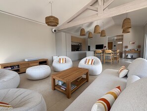 A modern living room with a sofa, ottoman, and coffee table, a kitchen in the background, and a dining area with wooden chairs. - MODERN VILLA WITH POOL (Châtelaillon-Plage)