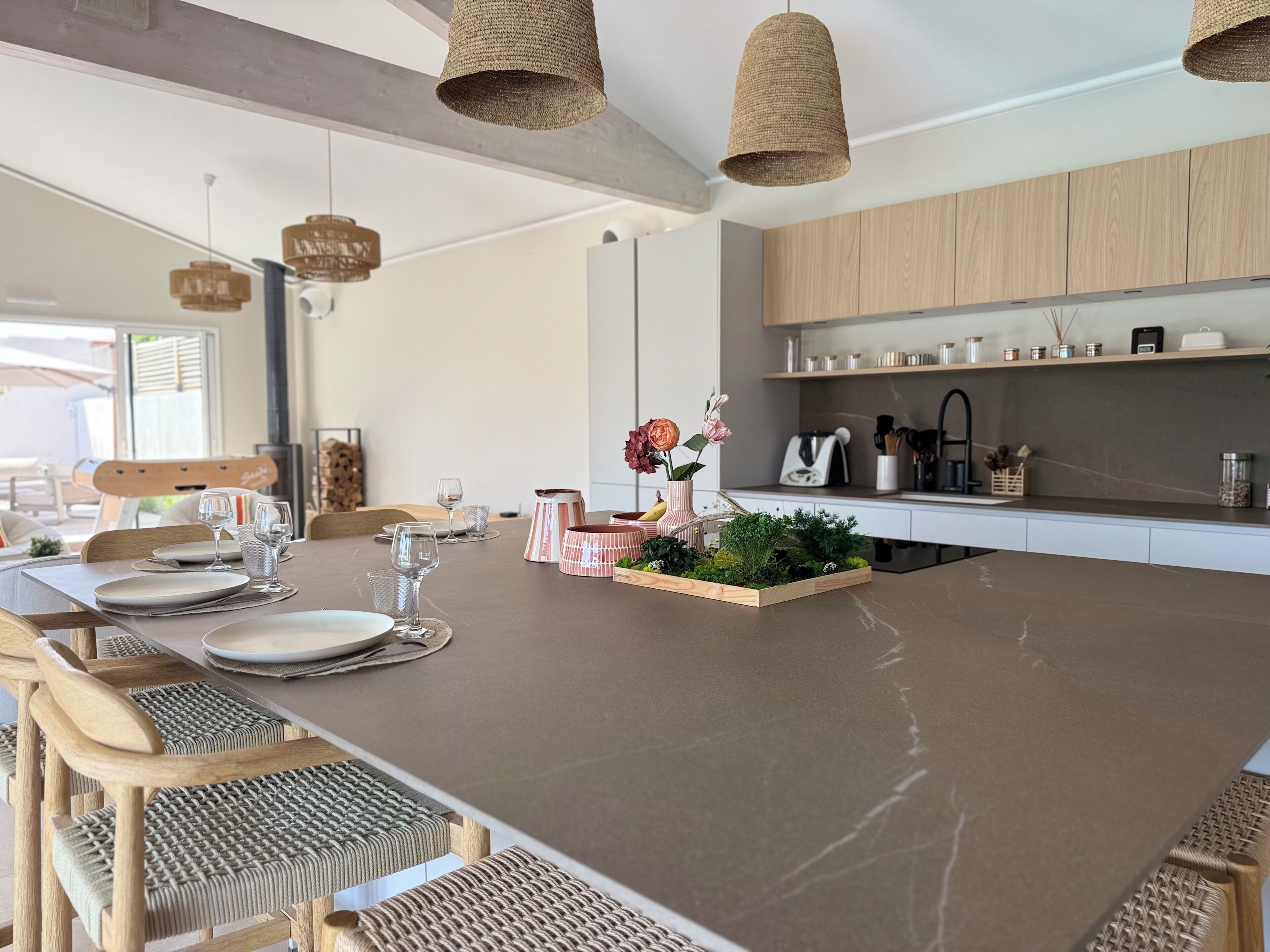 Une cuisine moderne avec des meubles en bois, un îlot central agrémenté d’une plante et des suspensions en osier.