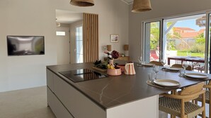 Moderni keittiö, jossa on keskeinen saareke, riippuvat, punotut valaisimet, litteänäyttöinen televisio ja näkymä ulos.