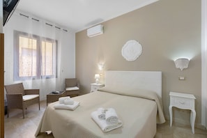 Hotellihuone, jossa on sänky, kaksi tuolia, yöpöytä, lamppu ja ikkuna, jossa on verhot.