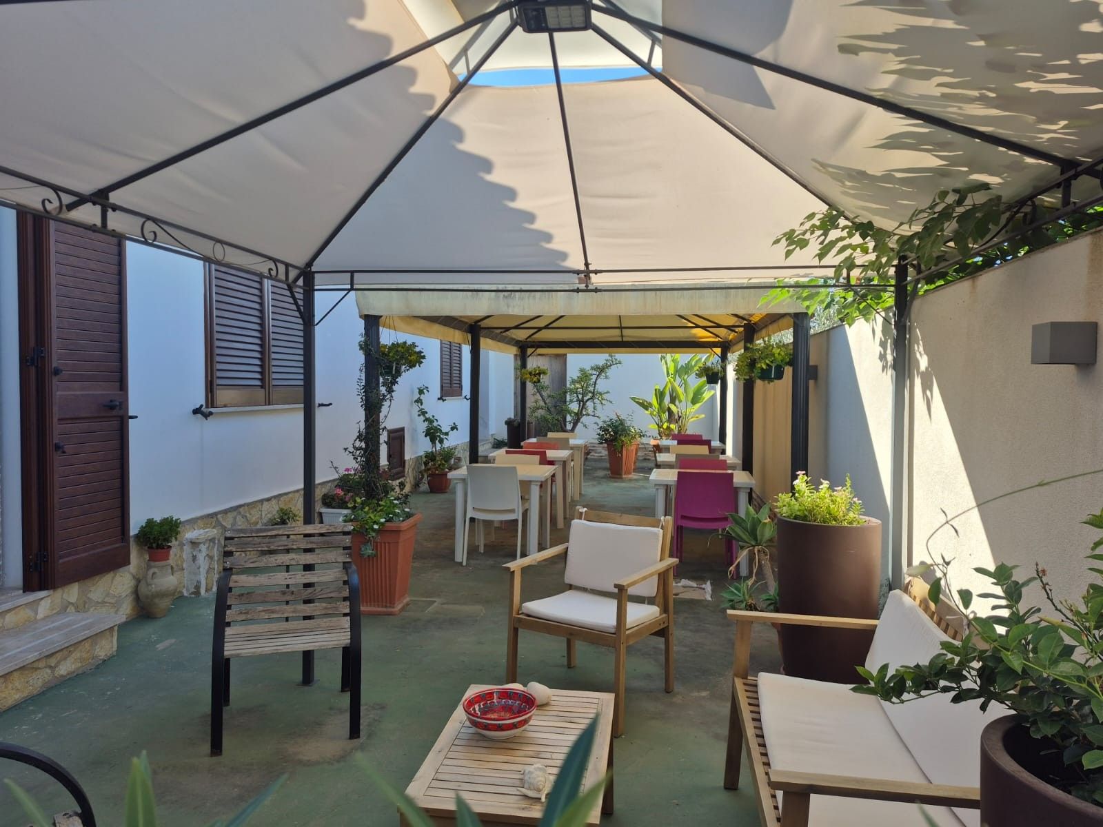 Un patio al aire libre con mesas, sillas, plantas en macetas y una sección cubierta.