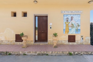 A building with a wooden door, two potted plants, a tiled mural, and a stone wall. - Room 'Demetra Malophoros - 16' with Shared Terrace, Wi-Fi and Air Conditioning (Marinella)